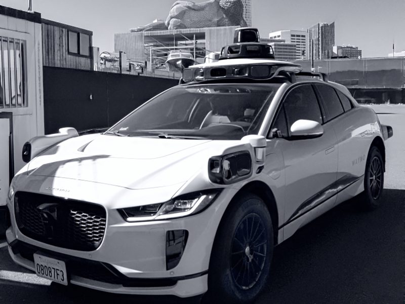 A white autonomous vehicle equipped with various sensors and cameras is parked outdoors. Urban buildings are visible in the background.