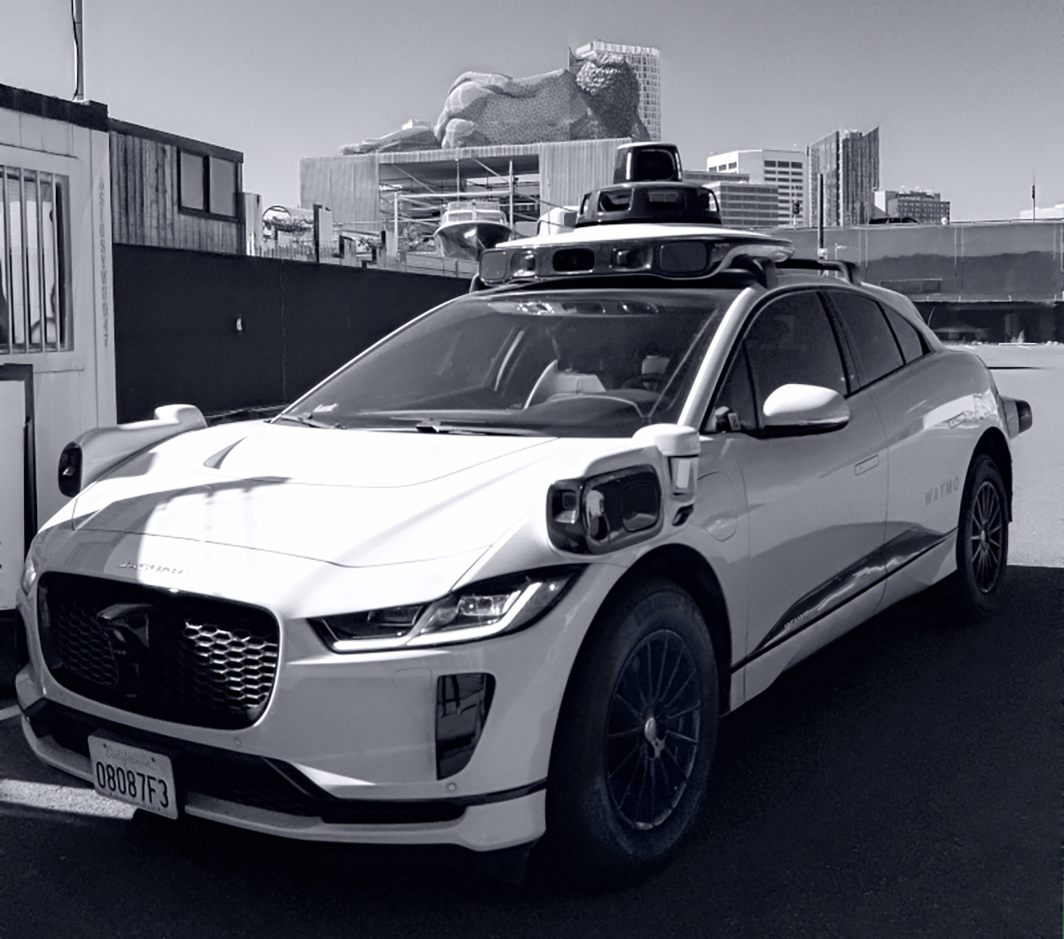 A white autonomous vehicle equipped with various sensors and cameras is parked outdoors. Urban buildings are visible in the background.