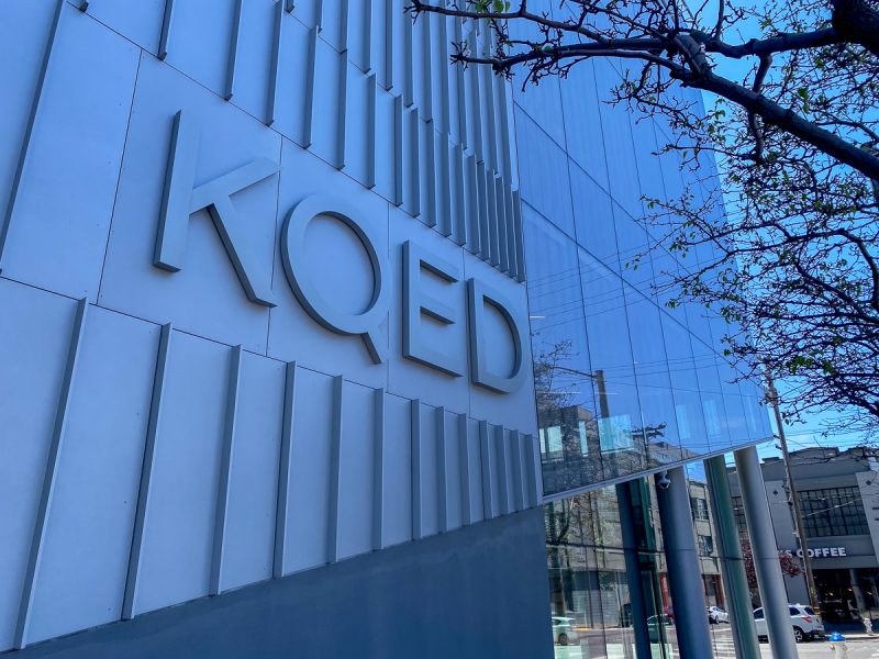 A building with large, white letters spelling "KQED" on its facade stands tall, glass exterior reflecting nearby structures and trees.