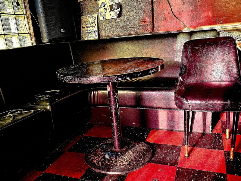 An empty vintage-style diner booth with a worn red leather seat, a single chair, and a round table, set on a black and red checkered floor.
