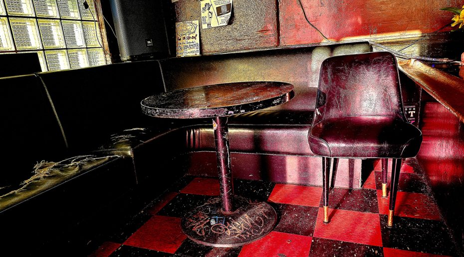 An empty vintage-style diner booth with a worn red leather seat, a single chair, and a round table, set on a black and red checkered floor.