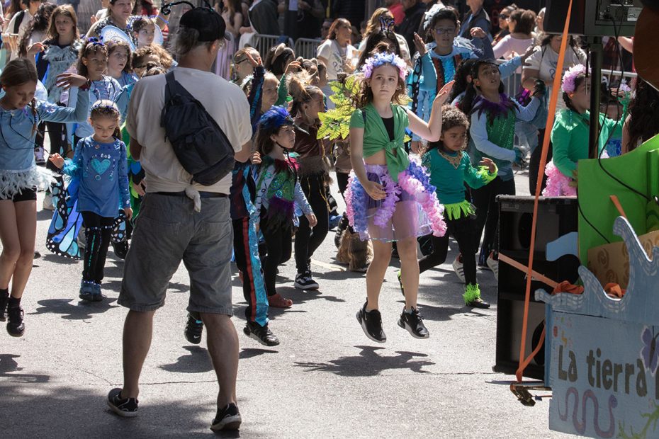 A group of children in colorful costumes dance in a parade while a man with a backpack watches from the foreground. A banner with "La tierra" is partially visible on the right.