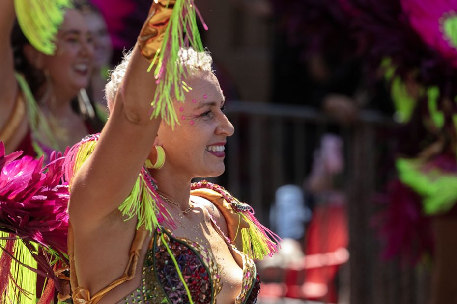A person in colorful, feathered attire smiles and raises their arms during an outdoor celebration or parade.