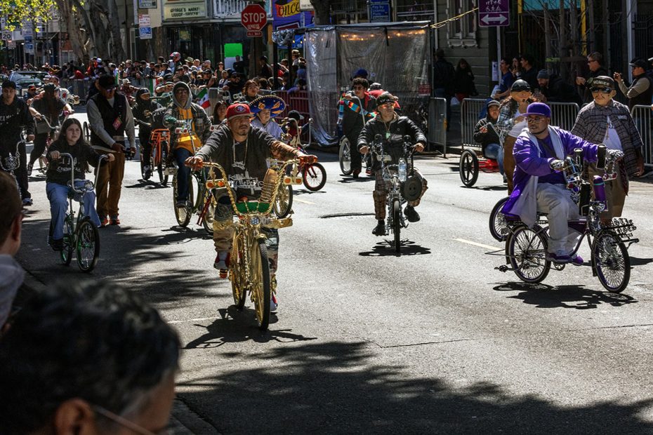 People riding decorated bicycles in a parade on a city street with onlookers behind barricades.
