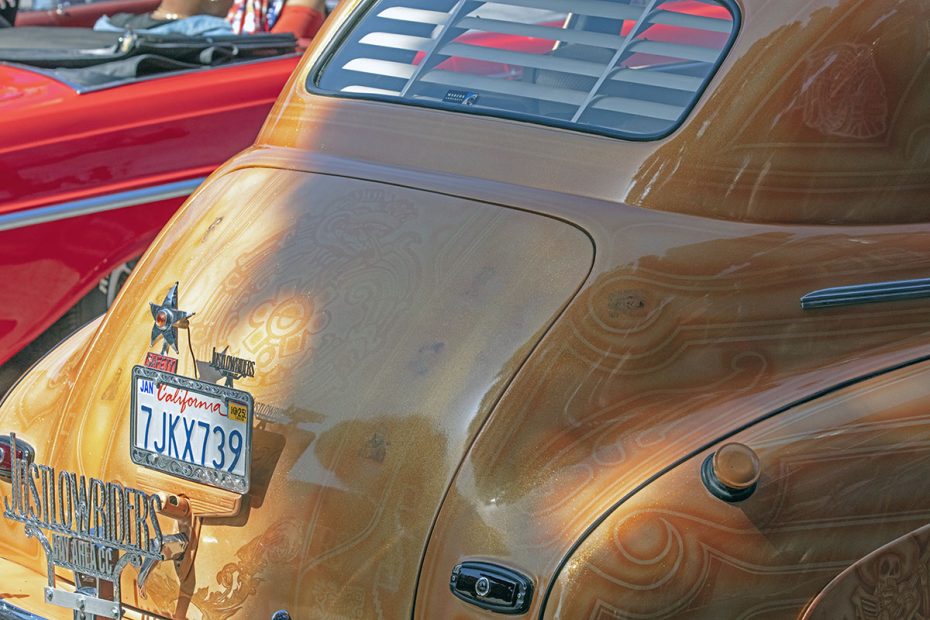 Rear view of a vintage car with intricate artwork on the trunk, California license plate 7JSK739, and visible louvers on the rear window, parked beside a red vehicle.