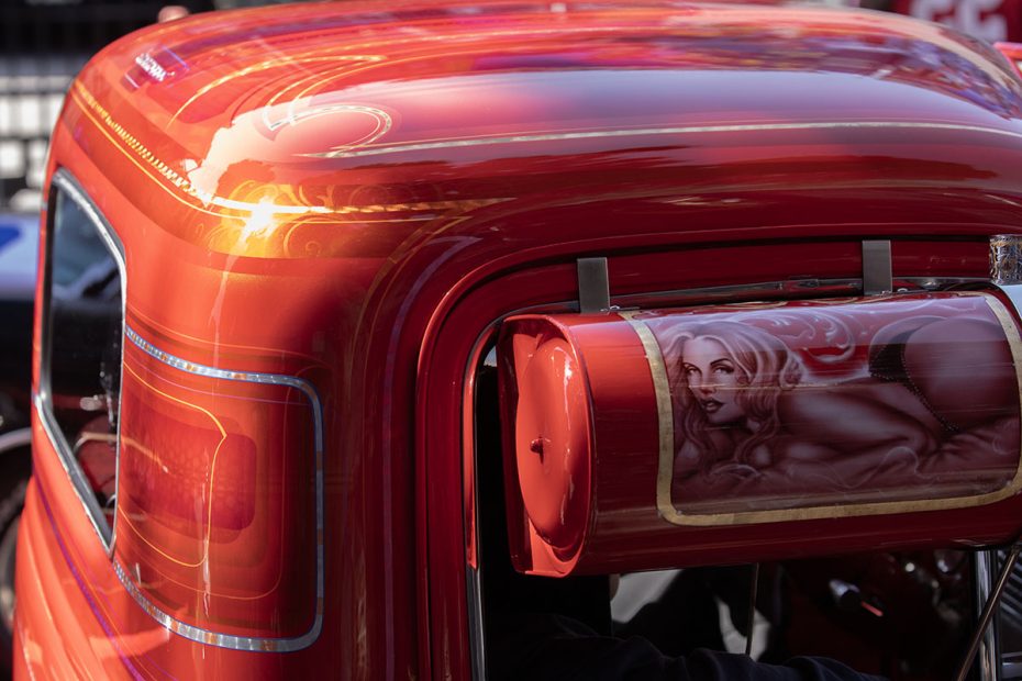 Close-up of a glossy red vehicle with intricate pinstriping and a painted image of a woman on the side of the rear part. Sunlight reflects off the shiny surface, highlighting the detailed artwork.