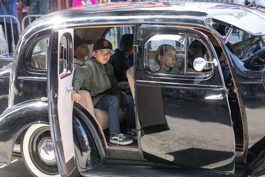 A group of children wearing baseball caps sit inside an open door of a classic black car, with one child looking forward.