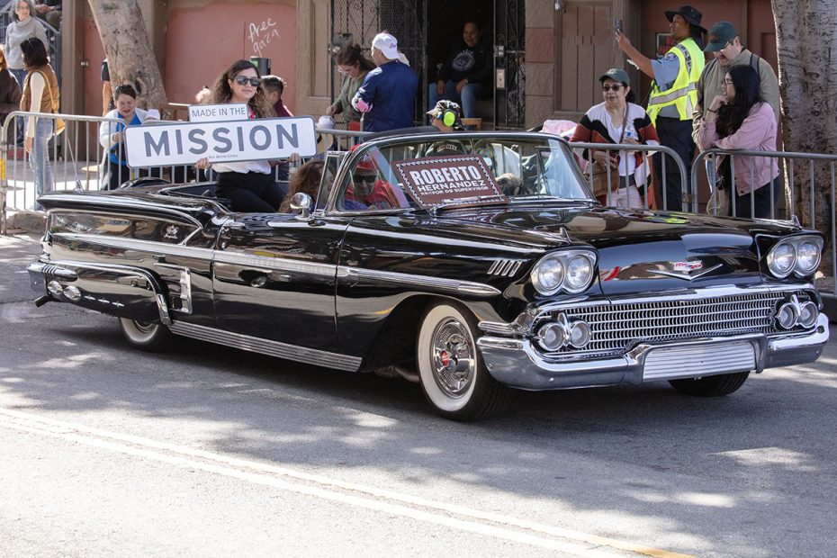 A black classic convertible car drives in a parade with a sign "Made in the Mission." A woman holds the sign, and people observe from the sidewalk, with a security guard in the background.