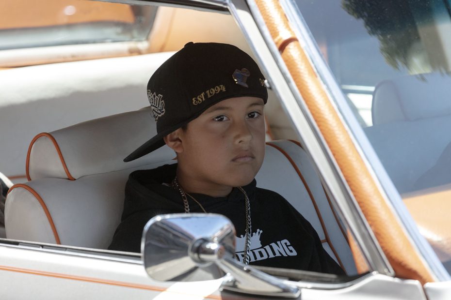 Young boy in black hoodie and hat sits in the driver's seat of a classic car, looking straight ahead through the open window.