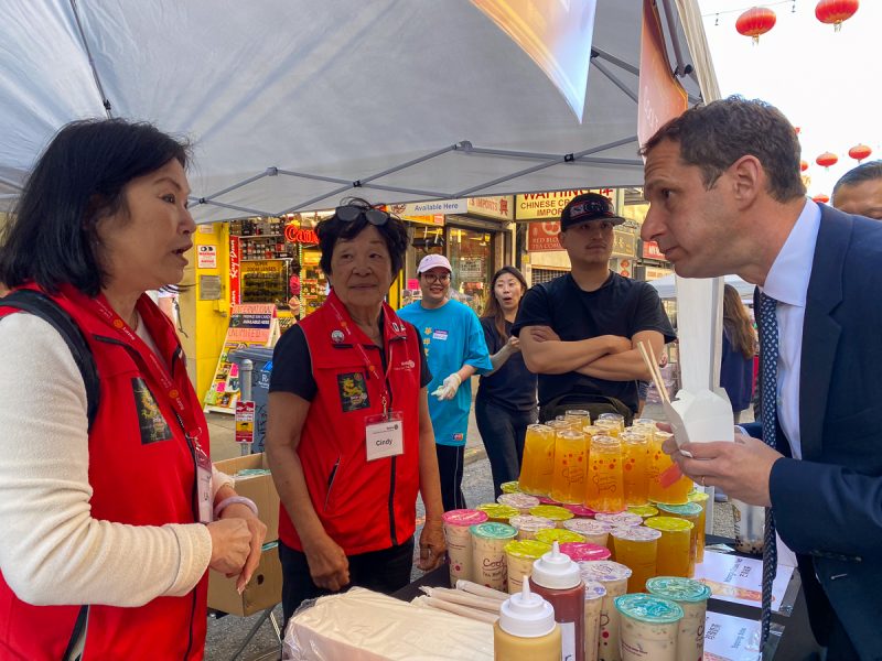 A man converses intently with a woman at a busy street market stall selling colorful beverages, with onlookers observing the interaction.