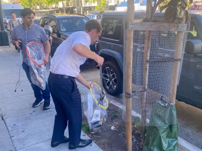 Two men on a city street, one in a dress shirt and tie collecting tennis rackets from a car trunk while the other watches.