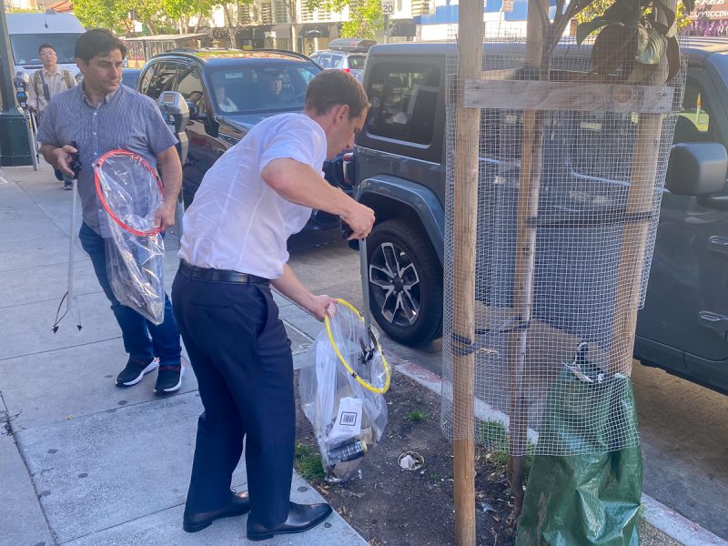 Two men on a city street, one in a dress shirt and tie collecting tennis rackets from a car trunk while the other watches.