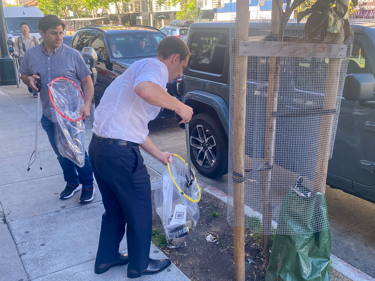 Two men on a city street, one in a dress shirt and tie collecting tennis rackets from a car trunk while the other watches.