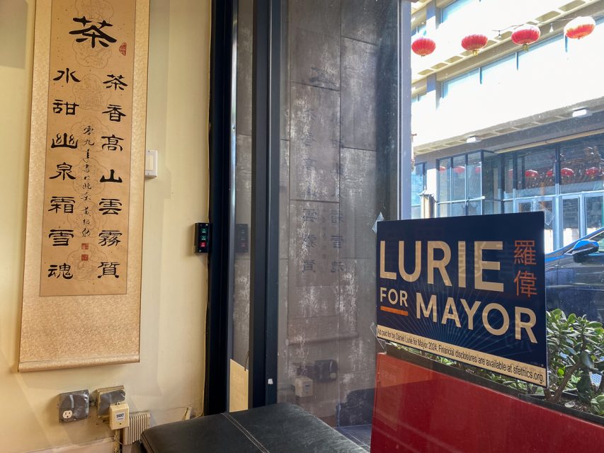 Interior of a building with a "lurie for mayor" sign, a decorative scroll with chinese calligraphy, and red lanterns visible through a window.