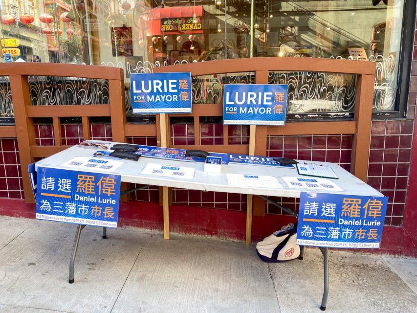 A campaign table with "Lurie for Mayor" signs and materials, some in English and others in Chinese, set up on a city sidewalk.