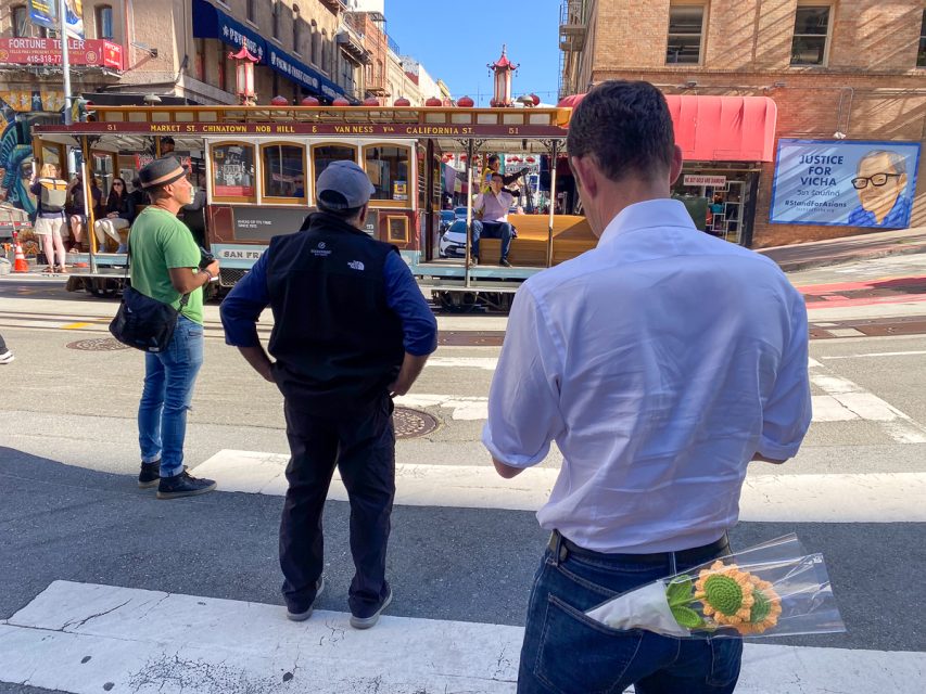 Three people standing on a city street, watching a cable car pass by; one man has two sunflower crochet in his backpocket.