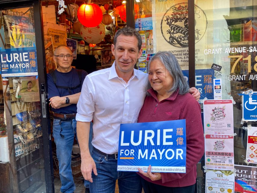 A man and a woman smiling, holding a "lurie for mayor" sign outside a shop decorated with red lanterns, while another man looks on from behind.