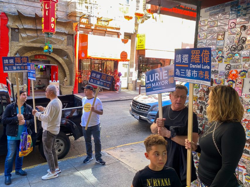 People campaigning for Daniel Lurie on a busy street in chinatown with various signs and cultural decorations.