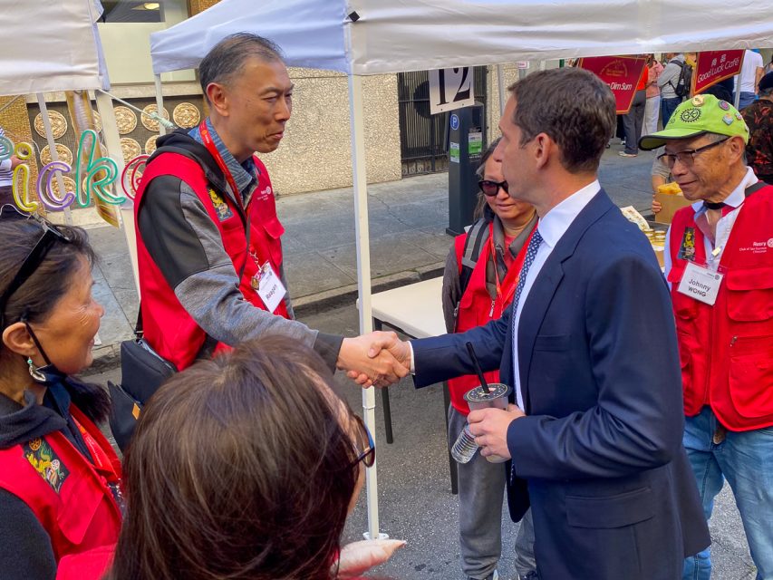 A man in a suit shaking hands with another man in a red vest at an outdoor event, surrounded by people also wearing red vests.