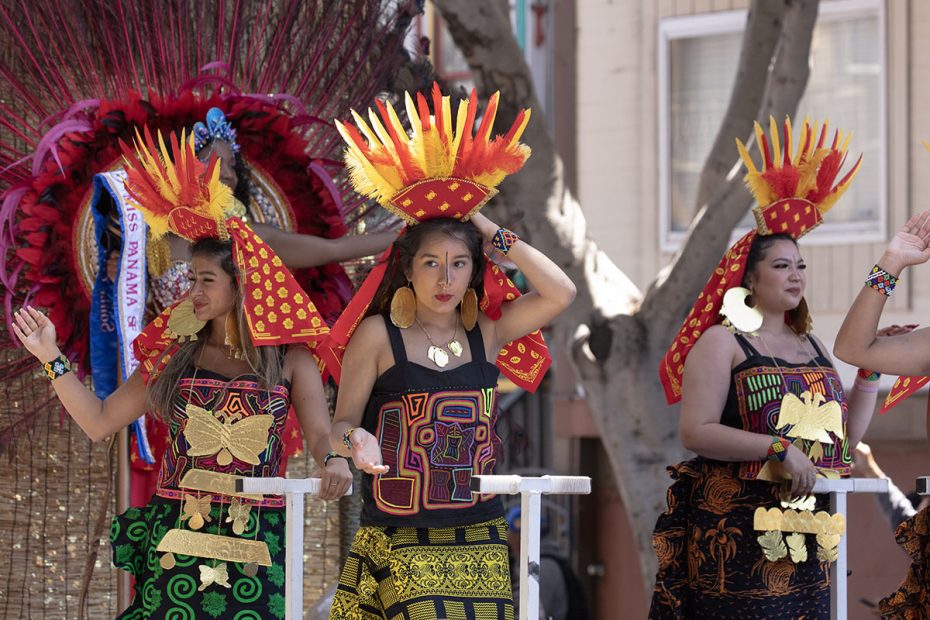 Three women in vibrant, traditional costumes with elaborate headpieces perform a dance outdoors, standing in front of a large decorative backdrop.