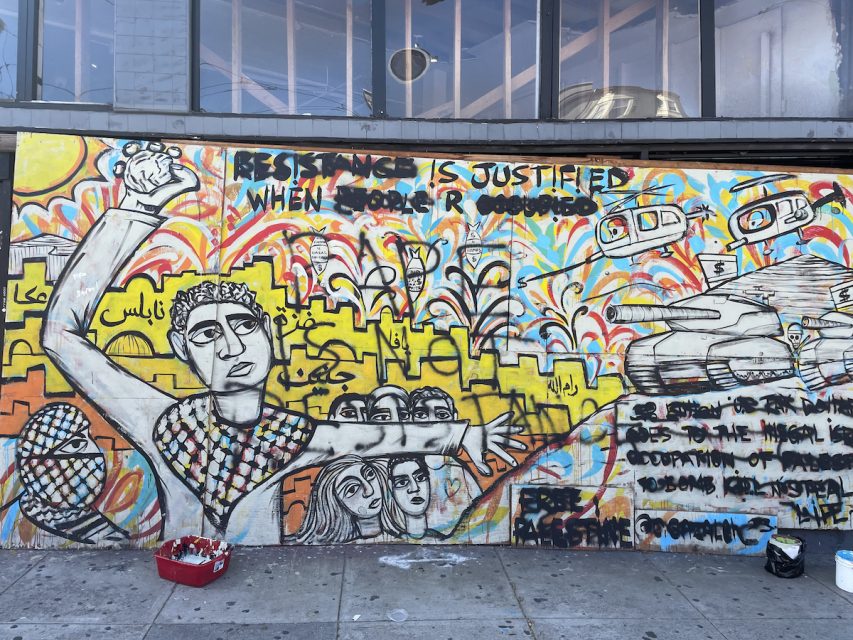 A vibrant mural depicts a figure with a raised fist in front of a cityscape with text reading "Resistance is justified when people are occupied," along with various protest symbols and messages.