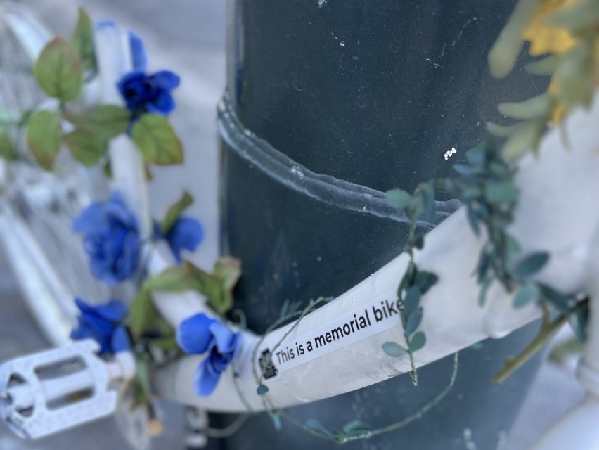 Close-up of a white memorial bike with blue flowers, attached to a pole, featuring a label that reads "This is a memorial bike.