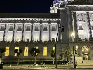 Night view of San Francisco City Hall illuminated, with a crescent moon visible above the building and a person on a bicycle in front.