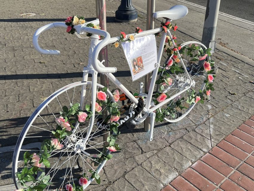 A white ghost bike adorned with floral tributes, chained to a street pole on an urban sidewalk, symbolizing a cyclist's memorial.