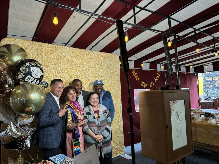 Group of five people standing together at a graduation celebration with a golden glitter backdrop. Balloons and decorations are visible in the background. A framed sign is on the right side of the image.