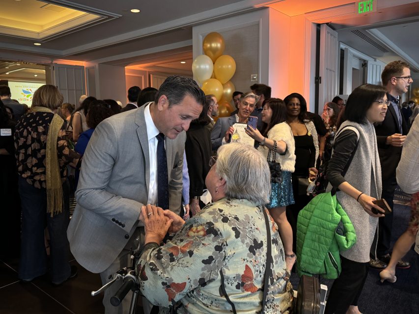 A man in a grey suit converses with an elderly woman using a walker at a crowded event; people are mingling in the background with balloons and warm lighting.