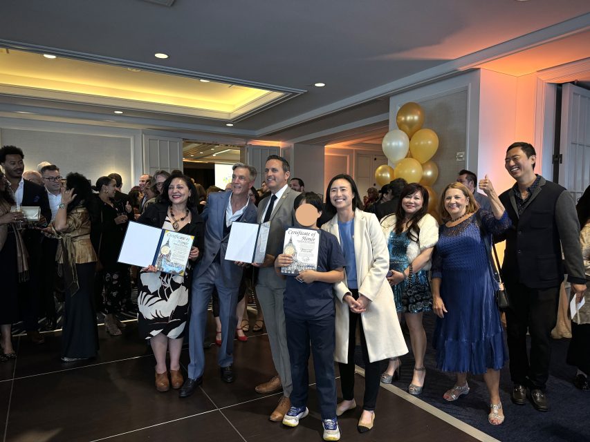 A group of people at an indoor event, with some holding certificates and posing for a photo. Others are mingling in the background. Balloons are visible to the right side of the image.