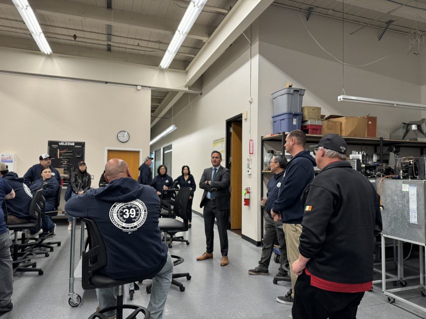A diverse group of workers in a workshop listening to a man in a suit speaking.