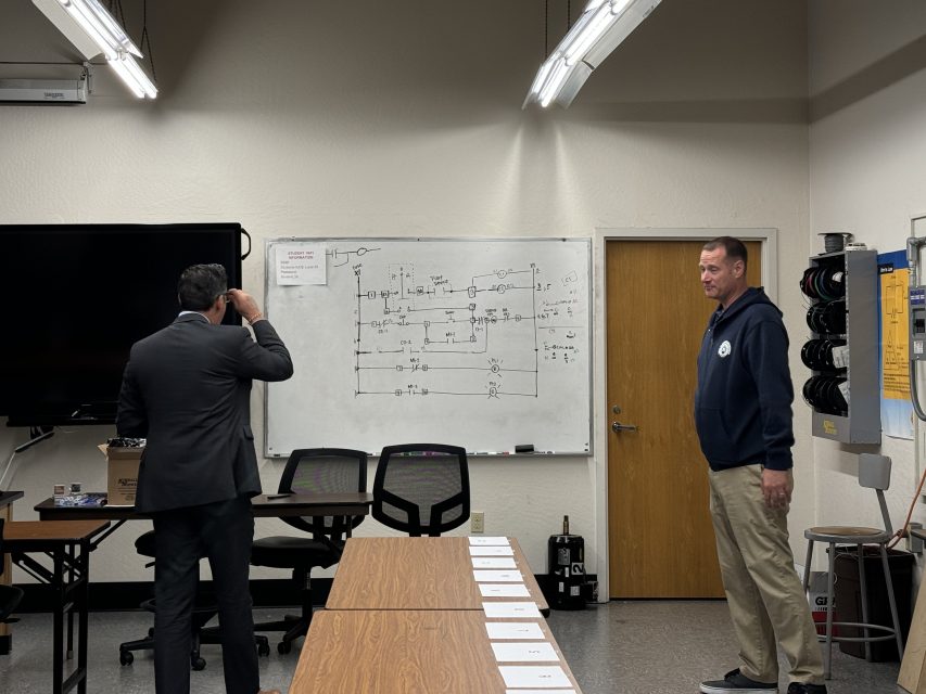 Two men in a classroom, one writing on a whiteboard and the other standing by, with diagrams displayed on the board and a TV screen on the side.