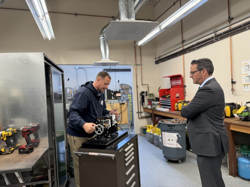 Two men in a workshop, one working on machinery components and the other observing, surrounded by tools and equipment.