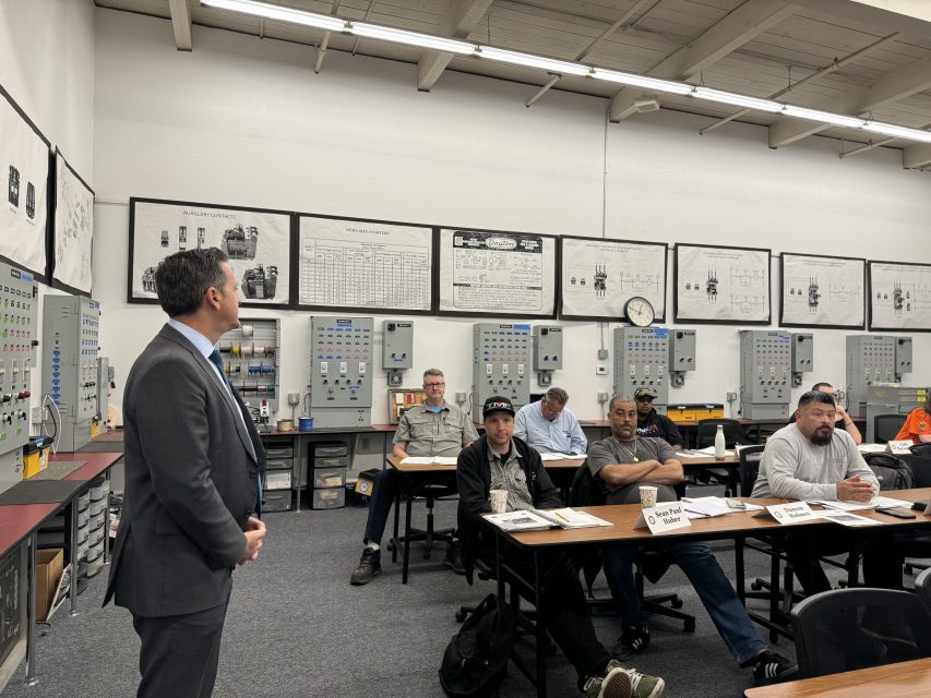 A man in a suit presents to a group of adult students in a vocational classroom filled with industrial equipment and instructional posters.