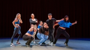 A group of seven people posing energetically on stage against a dark backdrop, dressed in casual activewear and performing dance poses.