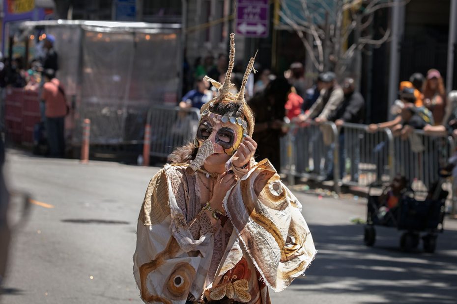 Person dressed in an elaborate costume with face paint and headpiece stands on a street during an outdoor event with spectators behind barriers.