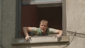 A person with short hair and a light-colored shirt leans out of a window, holding onto the window frame with both hands.