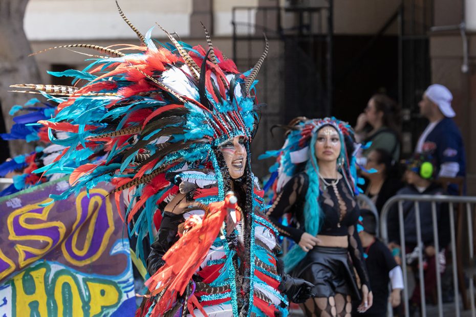 Two people in colorful feathered costumes participate in a parade. One leads with vibrant red, blue, and white feathers, while the other follows in matching colors with braided hair.
