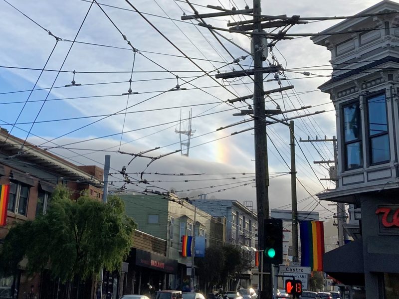 City street scene with tangled overhead wires, a rainbow fragment in the sky, and buildings displaying pride flags.