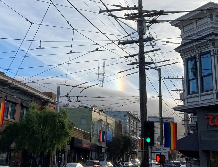 City street scene with tangled overhead wires, a rainbow fragment in the sky, and buildings displaying pride flags.