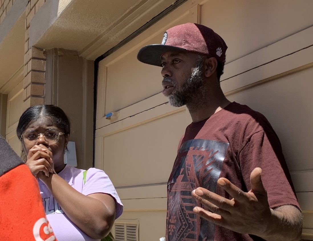 A man in a burgundy hat and graphic T-shirt gestures while standing next to a woman who appears concerned, holding her hand to her mouth. They are outdoors in front of a building.
