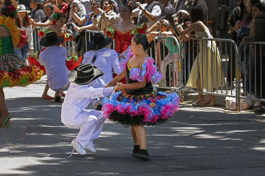Children in colorful traditional attire are dancing together in a lively outdoor event, with spectators watching behind a barricade.