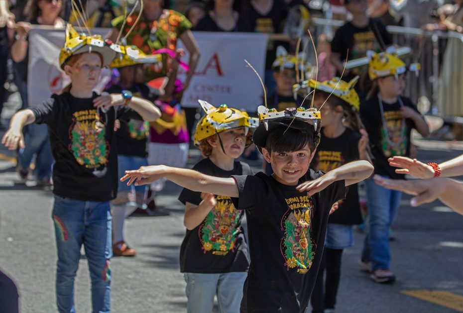 Children dressed in matching black t-shirts and yellow hats with decorations walk in a parade, arms outstretched, smiling as they perform.