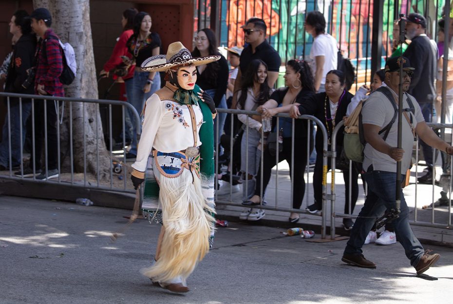 Person in traditional costume and large hat walking in a parade. Onlookers stand behind a barrier on the sidewalk.