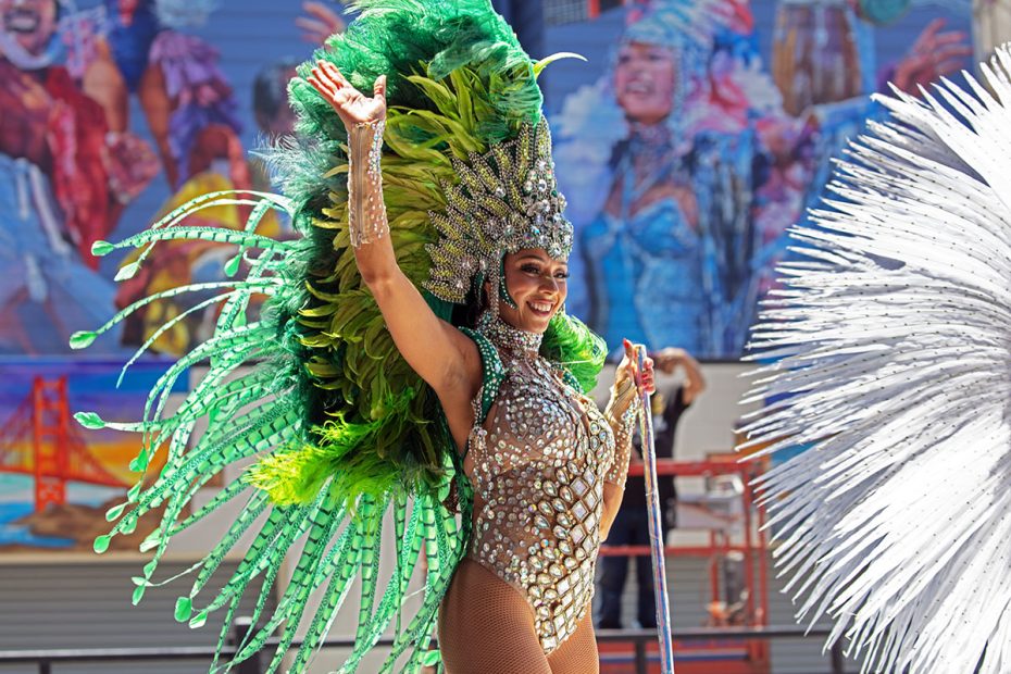 A dancer wearing a green feathered headdress and an embellished costume performs outdoors, with a colorful mural in the background.