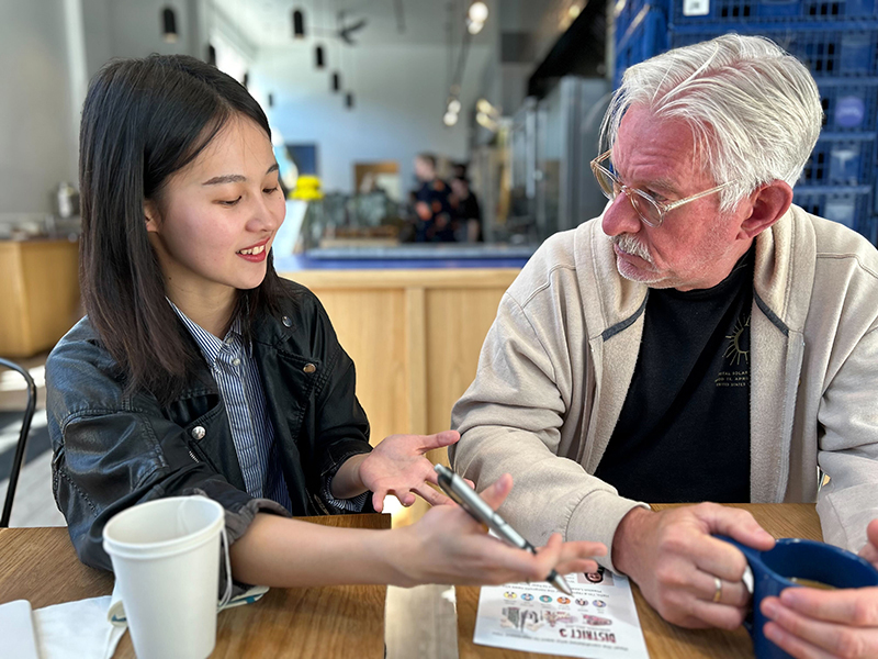 Young Asian woman showing smartphone to an older Caucasian man at a cafe table, both engaged in conversation.