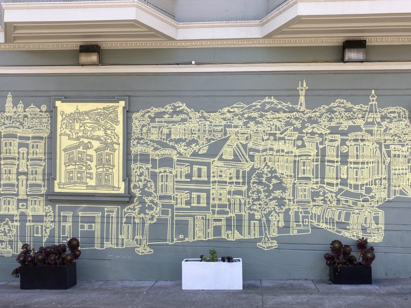 A wall mural with a yellow line drawing of a cityscape featuring detailed buildings, trees, and a prominent tower. Plants in black and white planters are placed in front.