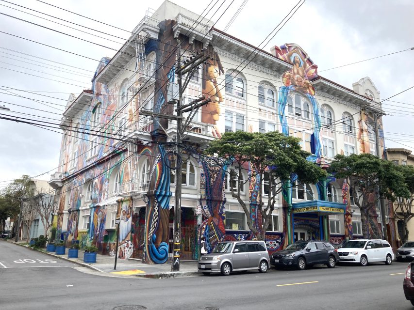 The four-story Women's building adorned with colorful murals stands at a street corner. Several cars are parked along the curb, and trees and power lines are visible in front of the building.