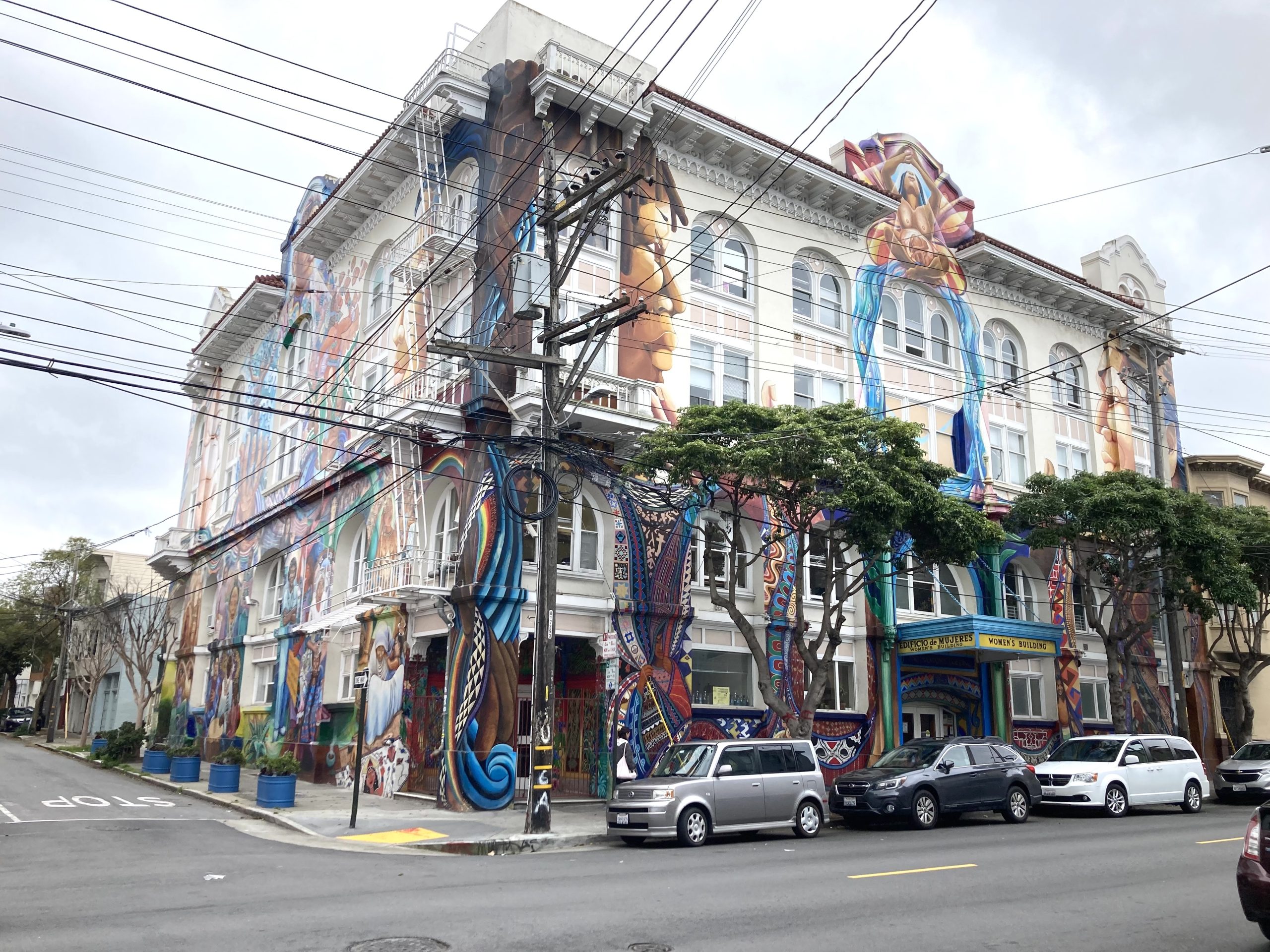The four-story Women's building adorned with colorful murals stands at a street corner. Several cars are parked along the curb, and trees and power lines are visible in front of the building.